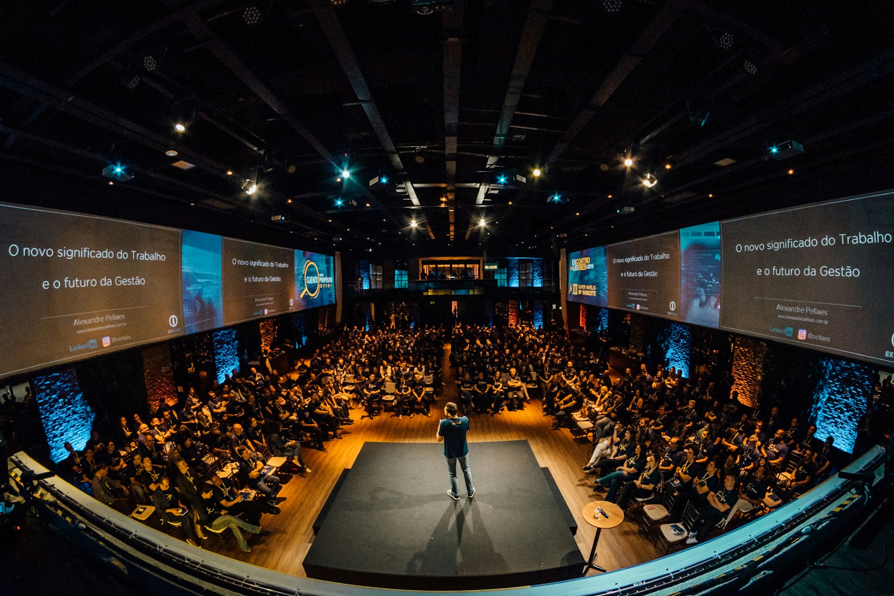 A full conference hall seen from above, a speaker on stage between two screens